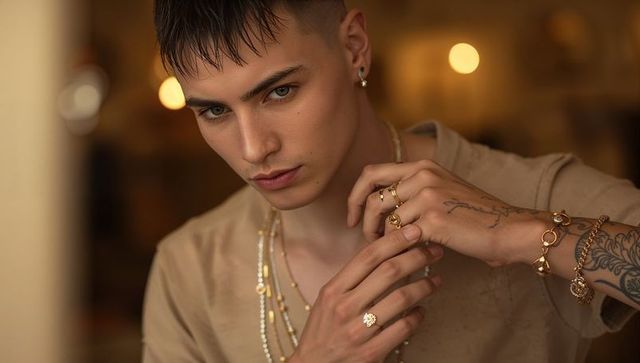 Stylish young man adjusting gold rings and layered necklaces in warm bokeh cafe portrait