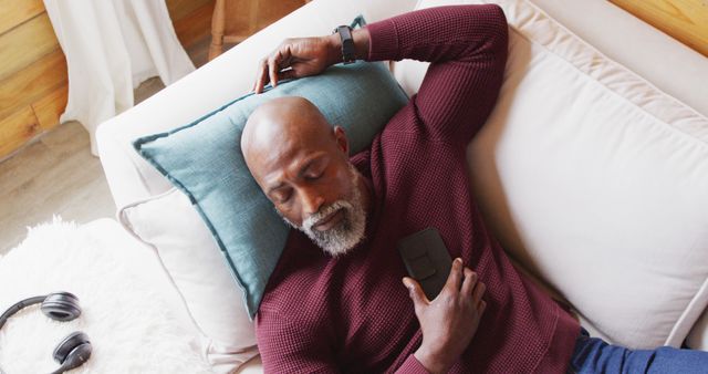 Mature bearded man in casual clothing relaxing on a couch, holding a smartphone. This image can be used for lifestyle or technology-related content, showing leisure, relaxation, and the use of mobile devices in everyday life.