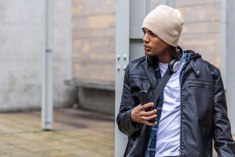 Young man leaning on column with smartphone and headphones in leather jacket and beanie