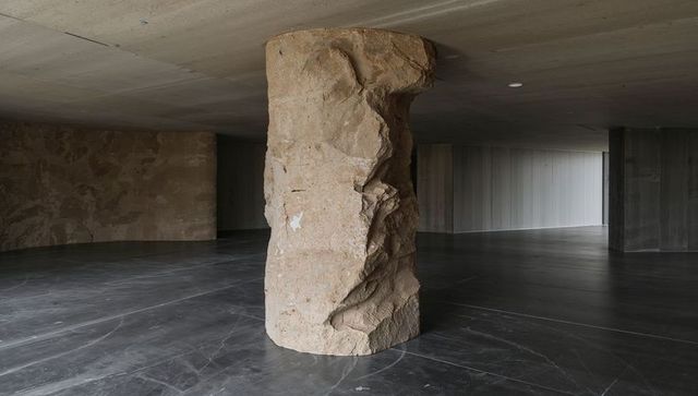 Rough stone column anchoring minimalist concrete gallery with polished dark marble floor