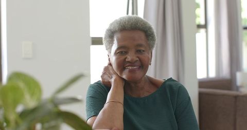 Senior african woman smiling while touching neck in bright living room