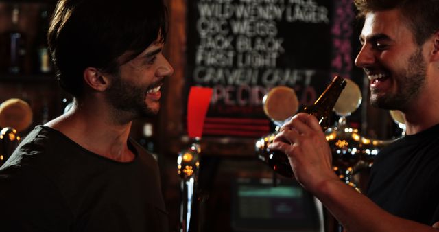 Two men enjoying a drink at a cozy bar, laughing and sharing a friendly toast with beer bottles. Perfect for use in campaigns about friendship, nightlife vibes, entertainment, and casual social gatherings. Also suitable for branding related to bars, pubs, and alcohol-related products and services.