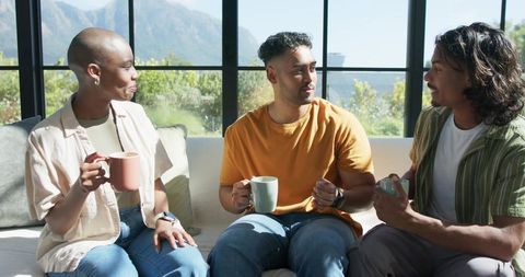 Friends chatting over coffee on sunlit sofa by large windows casual home conversation