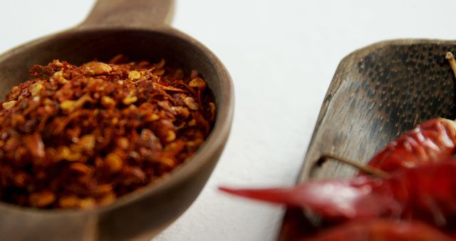 Ground red pepper in wooden spoon beside whole dry chili peppers on white surface. Perfect for use in culinary blogs, spice and seasoning websites, or food ingredient packaging and advertisements.