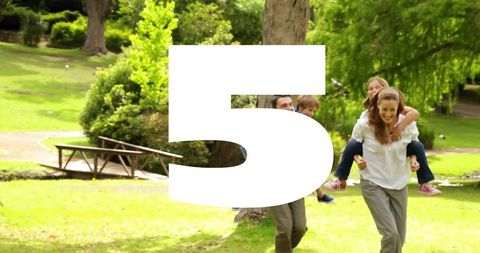Parents carrying children on piggyback walking across sunlit park lawn by wooden footbridge