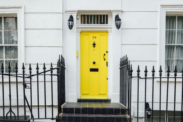 Bright yellow door stands out on classic white townhouse facade. Black iron fence frames entry. Use for urban living, exterior design ideas, residential listings, city lifestyle themes.