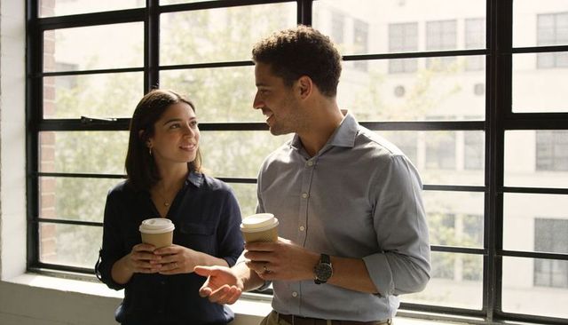 Colleagues sharing coffee and chatting by large industrial window in bright modern office