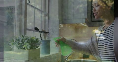 Woman Spraying Indoor Plant Near Kitchen Window with Green Sprayer