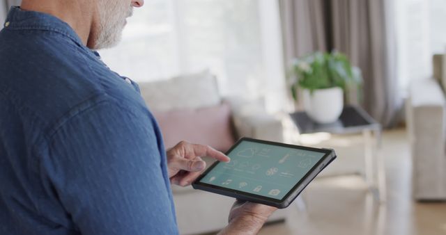 Senior man using a tablet to control smart home functions in a bright and modern living room. Shows touchscreen interface with multiple controls. Useful for representations of technology adoption in seniors, smart home technology, and modern living spaces.