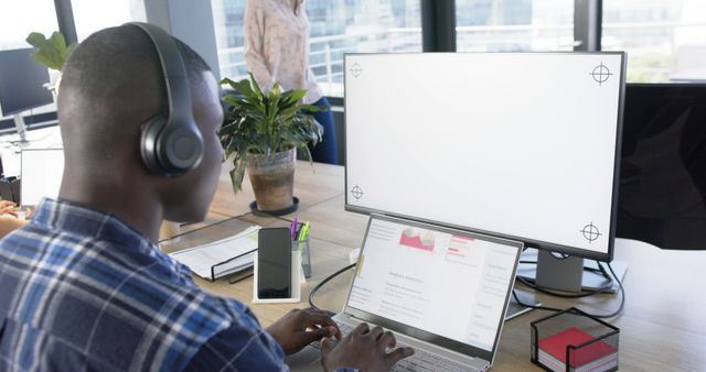 African American man focusing on work, wearing headphones, typing on laptop in modern office. Ideal for use in articles on remote work, technology, productivity tips, office cultures, and business environments.