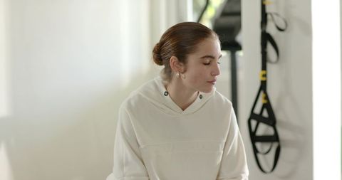 Young woman sitting in hoodie, gazing toward window by suspension trainer in home gym