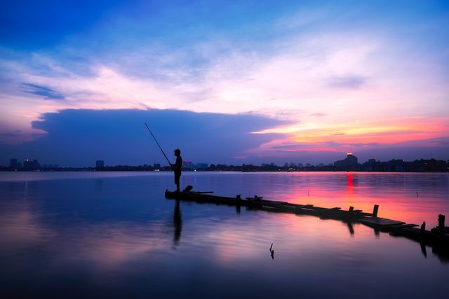 Silhouette of person fishing on a wooden pier during a stunning sunset over a calm lake. Perfect for themes related to fishing, tranquility, serene nature settings, outdoor activities, and beautiful twilight views. Ideal for use in blog posts, travel materials, relaxation and mindfulness content, and social media to showcase scenic and peaceful moments.