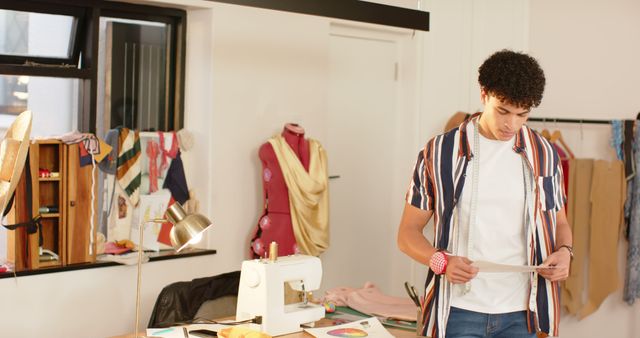 Young fashion designer reviewing pattern sheets in a vibrant craft studio. Ideal for use in articles about new fashion trends, creativity in fashion design, or DIY clothing projects.