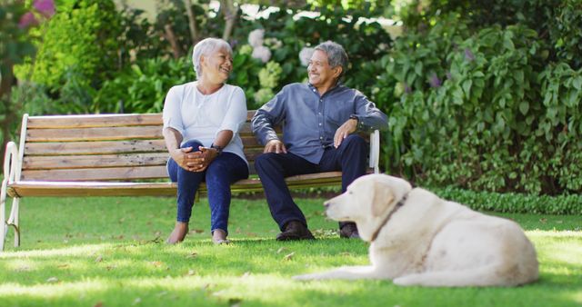 Image of happy biracial senior couple embracing and sitting on bench in garden with dog. active retirement lifestyle, senior relationship and spending time together concept.