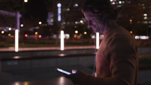 Depicts a Caucasian man wearing casual clothes and earphones, walking in a city at night, using his smartphone and holding a skateboard, indicating an active, connected urban lifestyle. Useful for topics related to city life, urban culture, digital technology, people on the move, and youth culture.