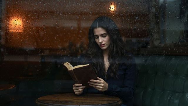 Woman reading novel in cozy rainy cafe window seat, moody intimate evening reading scene
