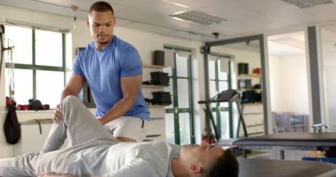 Trainer guiding assisted leg stretch on treatment table in sunlit fitness clinic