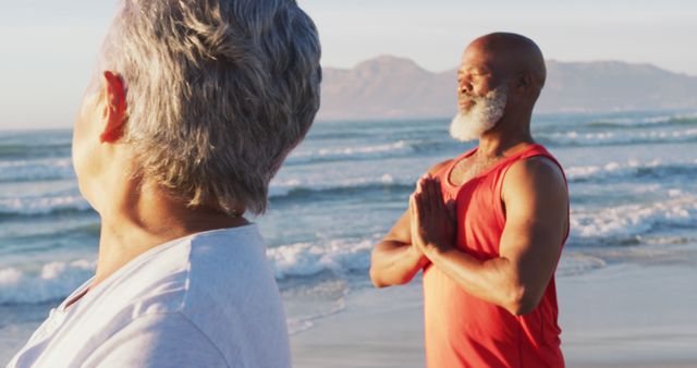 Senior man and woman meditating on beach at sunrise. Man practices yoga with hands in prayer pose, while woman turns head towards view. Ideal for promoting wellness, relaxation, serenity, physical and mental health in elderly, couple activities, or coastal travel destinations.