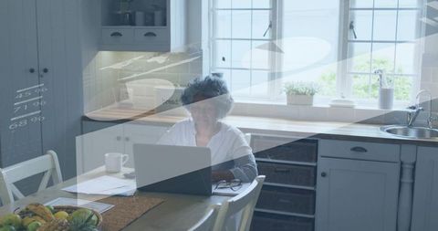 Senior woman working at kitchen table with laptop and documents, cozy home office daylight