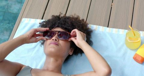 African american woman relaxing poolside adjusting sunglasses on towel with lemonade and sunscreen
