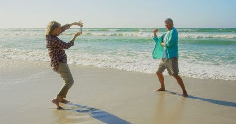 a senior man and woman enjoy playful photography at the beach. the woman takes photos with her mobile phone as the man strikes a pose. perfect for illustrating concepts of active retirement, joyful leisure time, or outdoor hobbies for seniors.