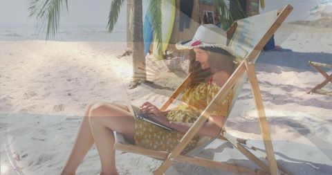 Woman working remotely on tropical beach in deck chair wearing straw hat and sundress