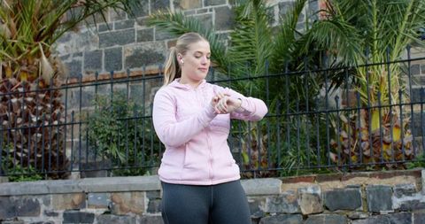 Checking smartwatch wearing earbuds in pink hoodie during outdoor urban walk by palms