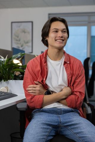 Young asian man sitting at desk in modern office wearing red shirt smiling confidently