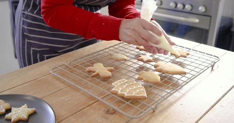 Woman in 30s decorating christmas cookies with icing piping bag on wooden kitchen counter