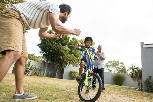 Father and grandfather encouraging young boy as he learns to ride a bicycle in the yard. Ideal for use in family-oriented advertisements, parenting blogs, outdoor activity promotions, and articles about childhood development and family bonding.