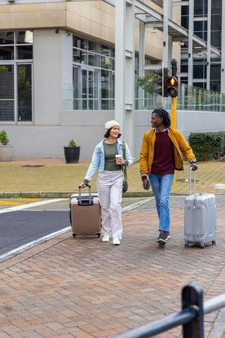Diverse couple walking with luggage on urban sidewalk pulling suitcases holding coffee