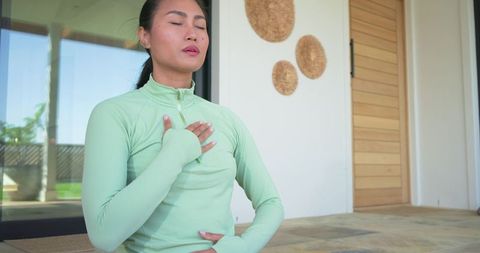 Asian woman practicing deep breathing meditation on sunny porch near glass door
