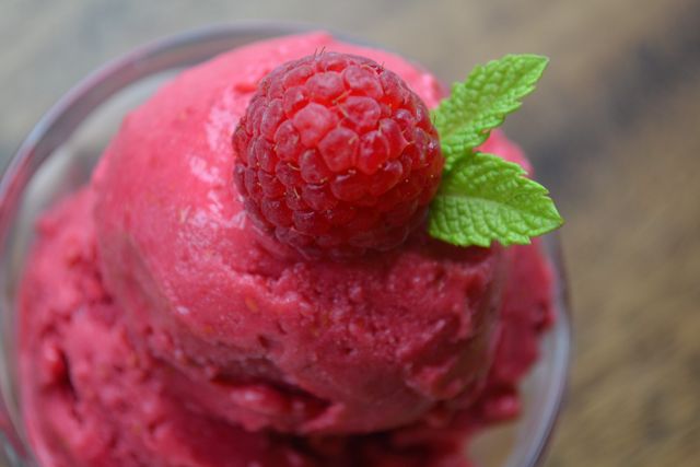 Close-up of a bright pink raspberry sorbet topped with a fresh raspberry and mint leaves, isolated on a wooden background. Perfect use for food blogs, restaurant menus, summer dessert promotions, and health food advertisements to highlight refreshing and healthy options during warm seasons.
