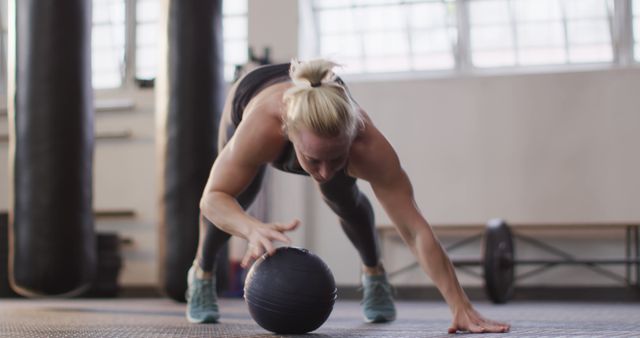 This image of a woman in a gym training with a slam ball on a mat is ideal for fitness blogs, gym promotions, or marketing materials for athletic wear. The intensity and focus depicted can inspire and motivate audiences to engage in physical activity and adopt a healthier lifestyle. It is also suitable for illustrating workout guides or fitness-related social media content.