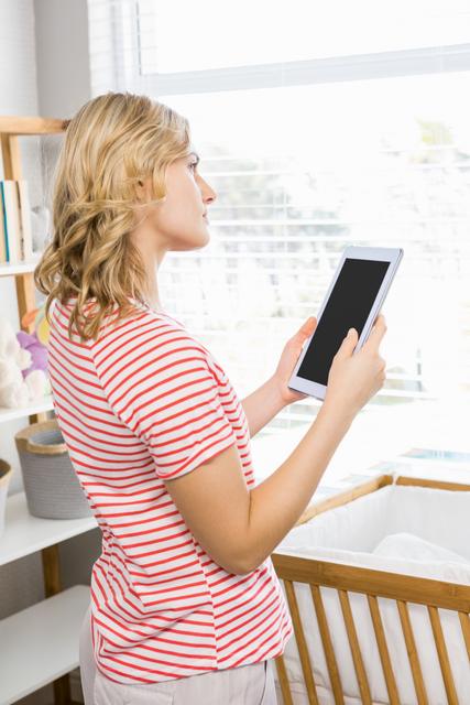 Young woman standing indoors holding a digital tablet, appearing thoughtful. She is wearing a casual striped shirt and standing near a window with natural light streaming in. This image can be used for themes related to technology, modern lifestyle, home environment, and contemplation.