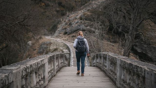 Female hiker crossing weathered stone bridge on rugged autumn trail, carrying pack and pole