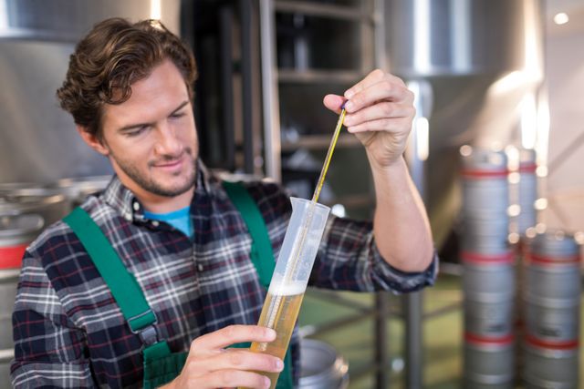 Worker in a brewery warehouse examining beer quality using a hydrometer. Ideal for use in articles or advertisements related to the brewing industry, quality control processes, or professional brewing equipment. Suitable for illustrating concepts of beer production, industrial work, and beverage testing.