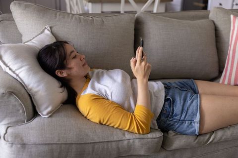 Asian woman lounging on grey sofa using smartphone for browsing and relaxation at home