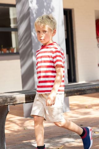Blond boy walking past weathered bench in sunlit courtyard wearing striped tee