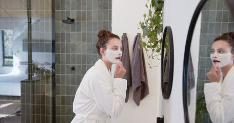 Woman in Bathrobe Applying Facial Mask in Modern Bathroom