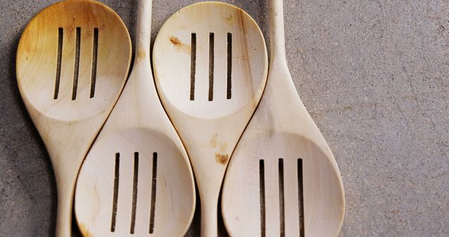 Three wooden slotted spoons are laid out on a concrete surface, with copy space. Their simple design suggests a rustic or homemade cooking theme, emphasizing the beauty of kitchen essentials.