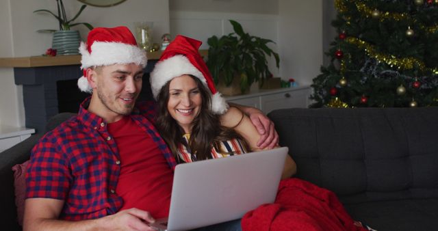Couple wearing Santa hats sitting on couch, video chatting on laptop. Christmas tree and fireplace in background. Ideal for use in holiday greeting cards, festive advertisements, or content related to remote communication during Christmas season.