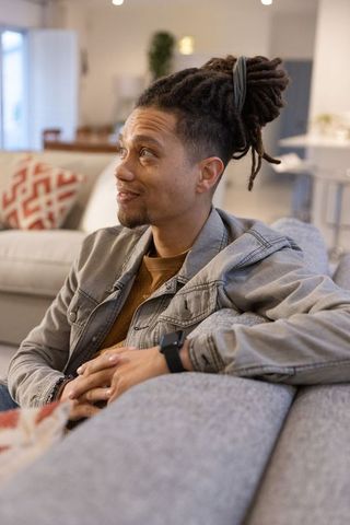 Relaxed young man with dreadlocks sitting on sofa wearing denim jacket and smartwatch