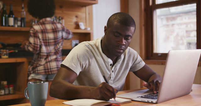 African american man using laptop at kitchen table and writing notes, copy space. Remote working, communication, city living, domestic life and lifestyle, unaltered.
