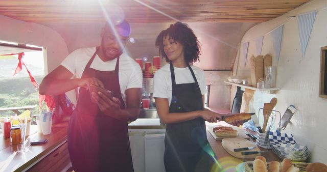 African american couple wearing aprons using digital tablet and preparing hot dogs in the food truck. food truck and street food concept