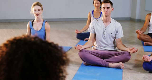 Group of young adults sitting on yoga mats in a serene setting, each participant practicing mindfulness and yoga. The class appears calm, focused on relaxation and stress relief. Perfect for promoting health benefits of yoga and meditation, wellness programs, fitness classes, and mindfulness retreats.