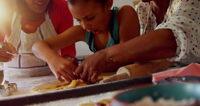 A young child, along with two adults, are engaged in baking in a kitchen. They are working together to cut dough, suggesting a hands-on learning experience and family bonding activity. The warm lighting emphasizes the cozy, familial atmosphere. This photo can be used for illustrating family bonds, home cooking, tradition, or educational activities for children.