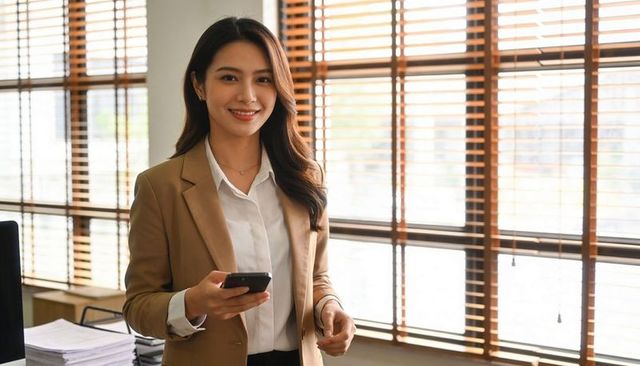 Smiling businesswoman holding smartphone in modern office with sunlit wooden blinds