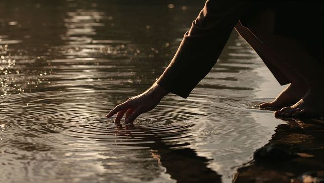 Solitary hand touching water ripples at twilight on rocky shore with bare feet reflection