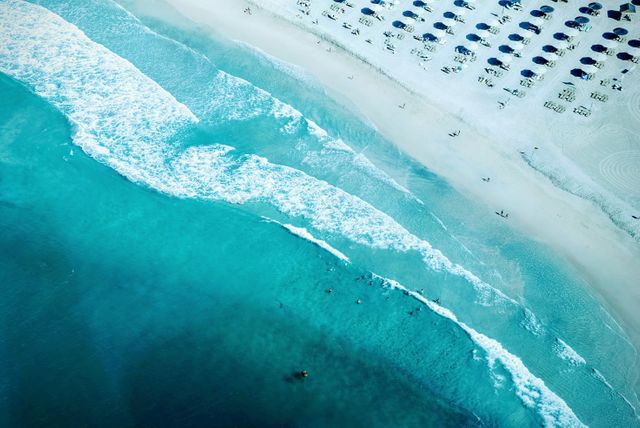 Aerial photograph capturing the vibrant blue waves of the ocean hitting the sandy shoreline. Rows of beach chairs with umbrellas along the beach highlight the area's inviting and relaxing atmosphere. Perfect for use in travel advertisements, tropical destination promotions, beach holiday themes, and nature-related content.
